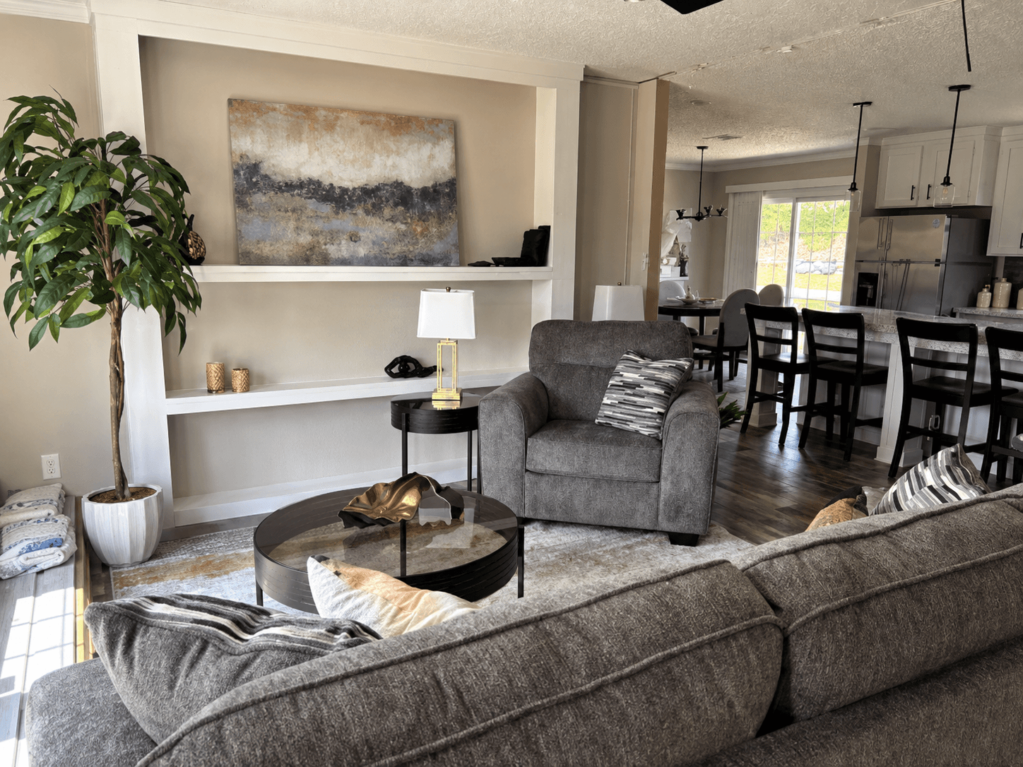 Open living room and kitchen with a grey sectional, glass coffee table, white built-ins and black barstools