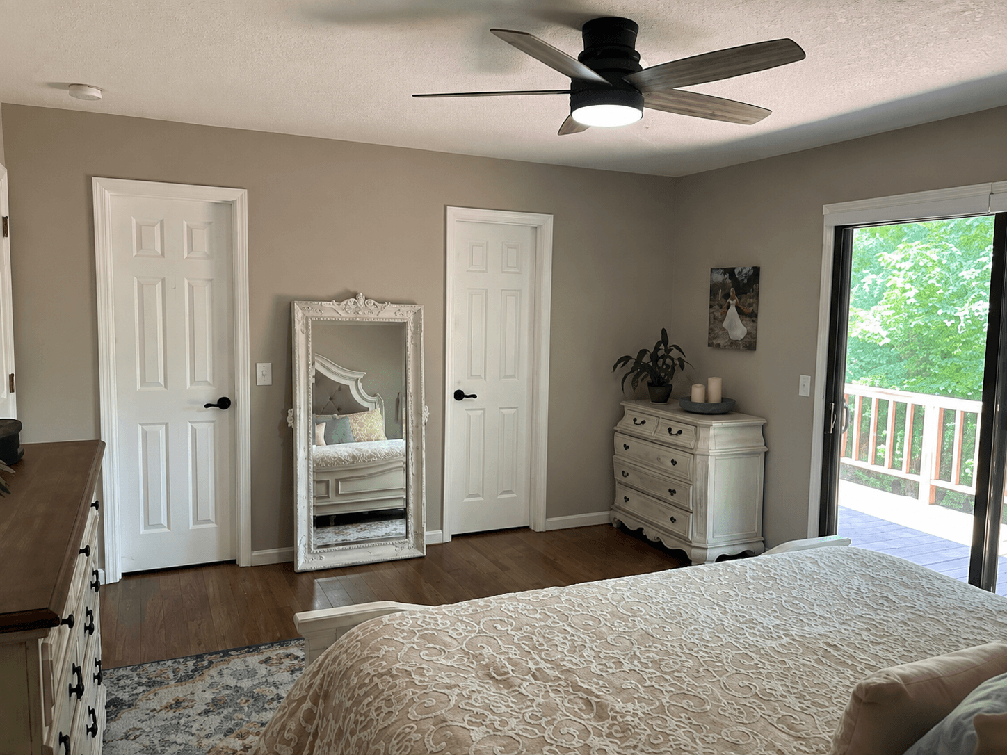 Furnished bedroom with greige walls, a cream quilt, a leaning floor mirror and a sliding door to the woods