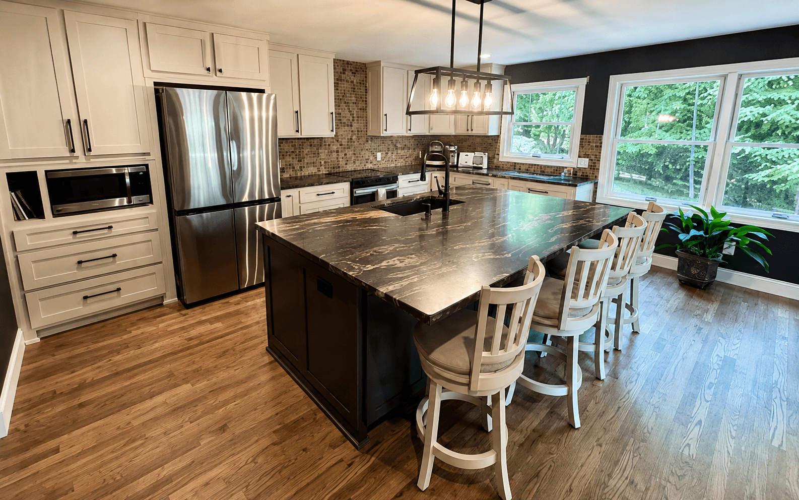 Kitchen island with a dark veined stone top, white barstools, a linear chandelier and a stainless refrigerator