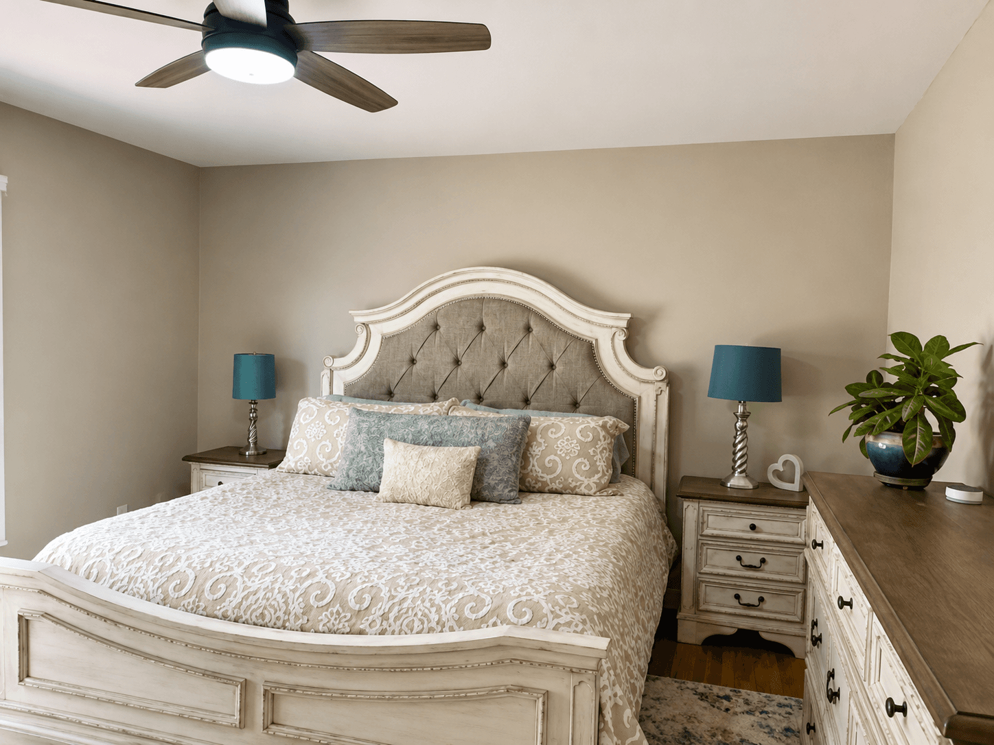 Bedroom with a tufted cream headboard, white nightstands, teal lampshades and a leafy plant
