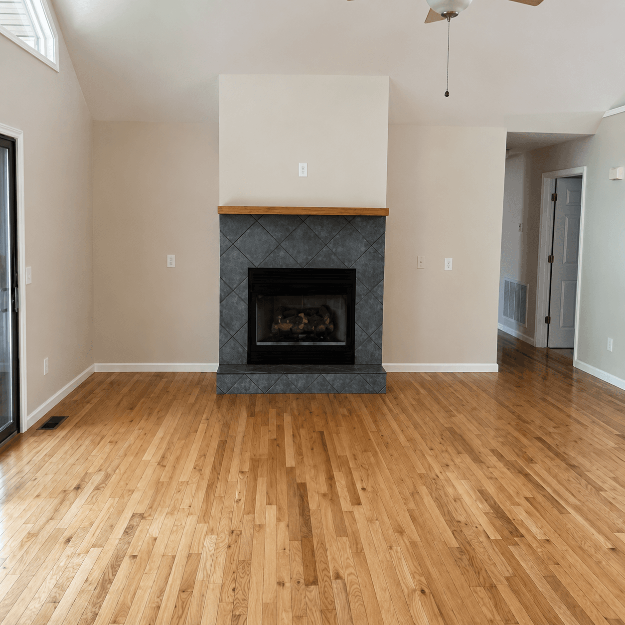 Empty living room with a tiled fireplace, wood mantel, vaulted ceiling and a sliding glass door