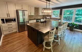 Kitchen island with a dark veined stone top, white barstools, a linear chandelier and a stainless refrigerator