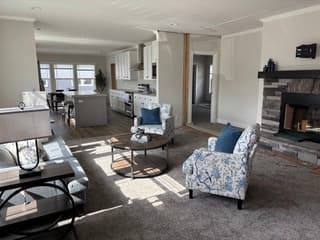 Open living room and kitchen with floral chairs, a stone fireplace and white shaker cabinets beyond