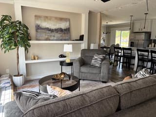 Open living room and kitchen with a grey sectional, glass coffee table, white built-ins and black barstools